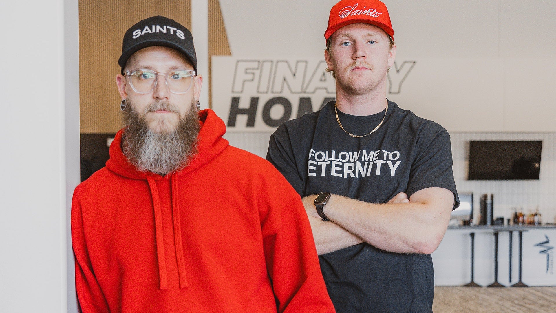 Two men standing in a room with a 'Finally Home' sign in the background