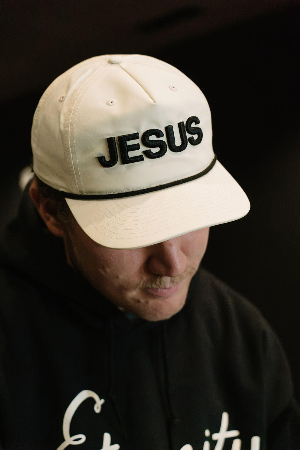 A man wearing a cream-colored baseball cap with 'JESUS' embroidered in black across the front, paired with a black hoodie against a dark background.